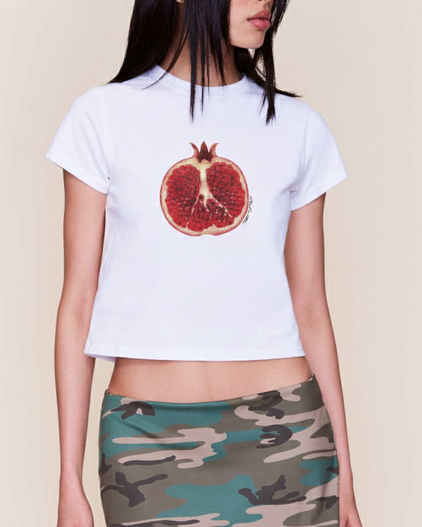 Person wearing a white t-shirt with a pomegranate graphic and a camouflage skirt on a beige background