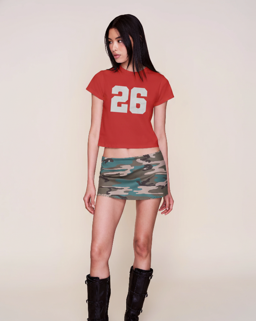 Person wearing a red t-shirt with the number 26 and a camouflage skirt on a plain background