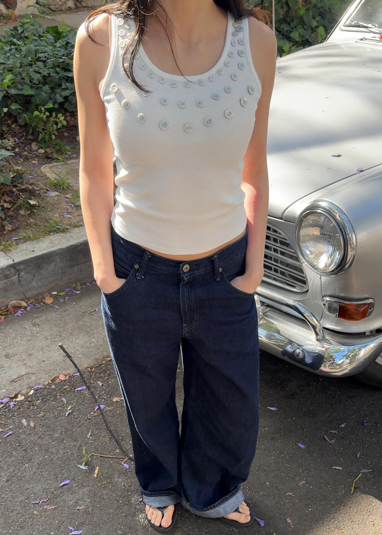 Person wearing a white tank top and dark blue jeans standing on a street with a vintage car in the background.