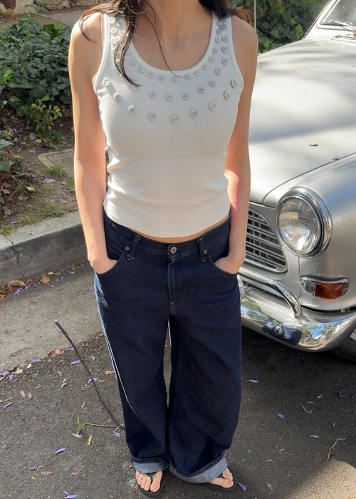 Person wearing a white tank top and dark blue jeans standing on a street with a vintage car in the background.