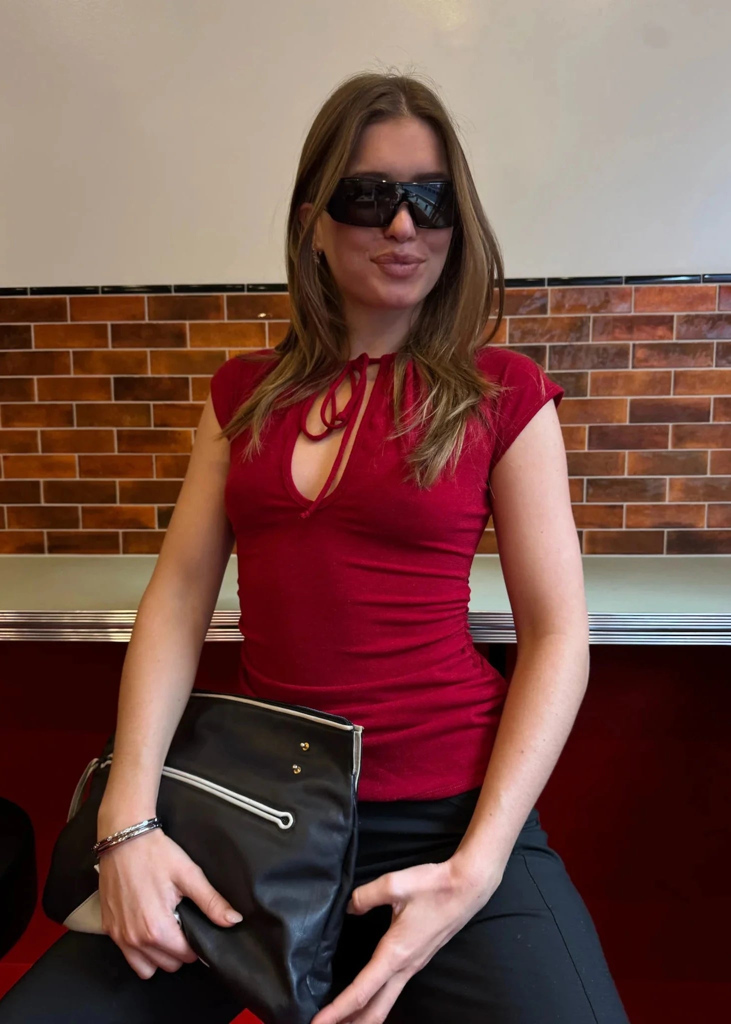 Woman in a red top and sunglasses holding a black clutch against a tiled wall.