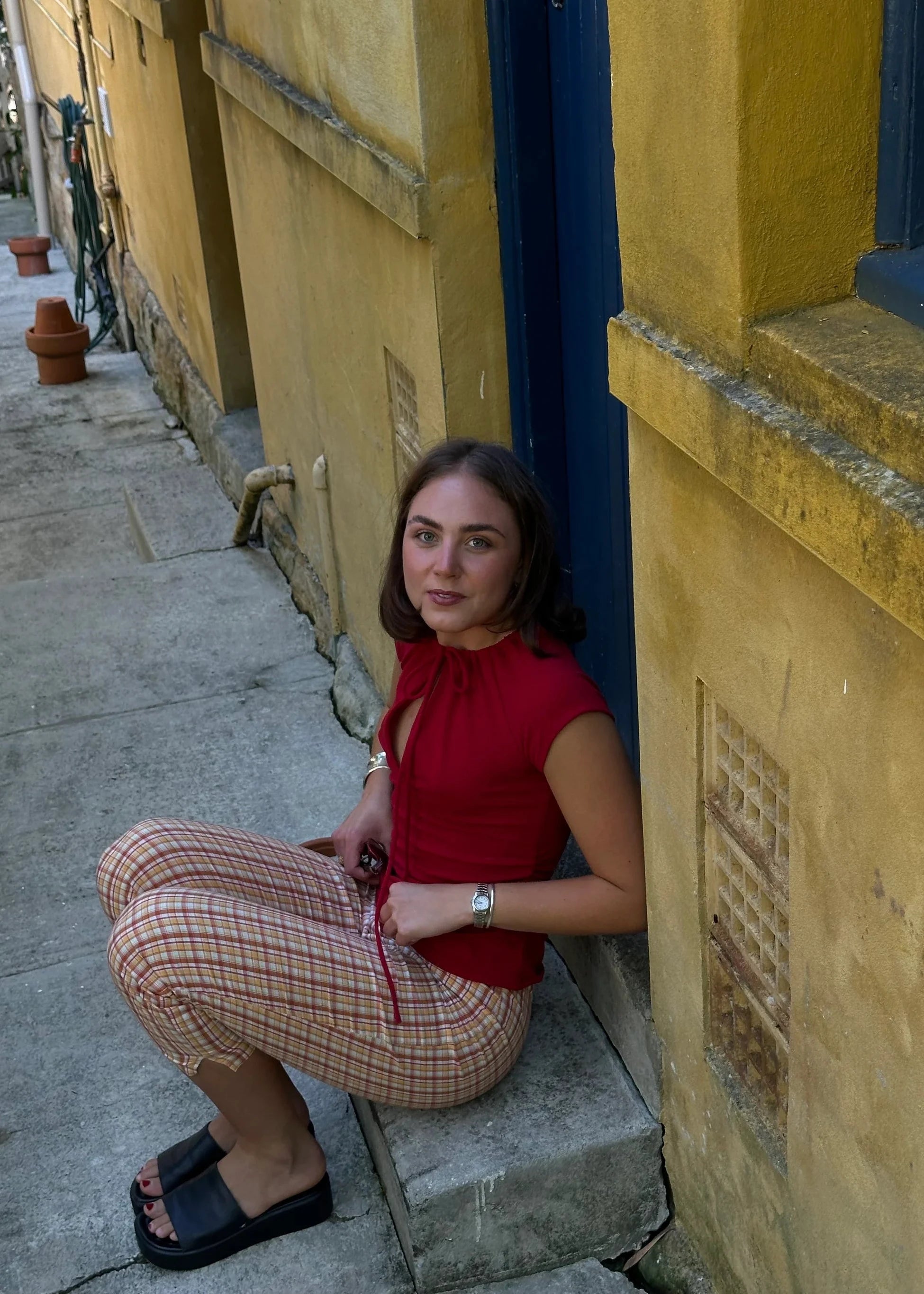 Woman in a red top and plaid pants sitting on a step next to a yellow wall.
