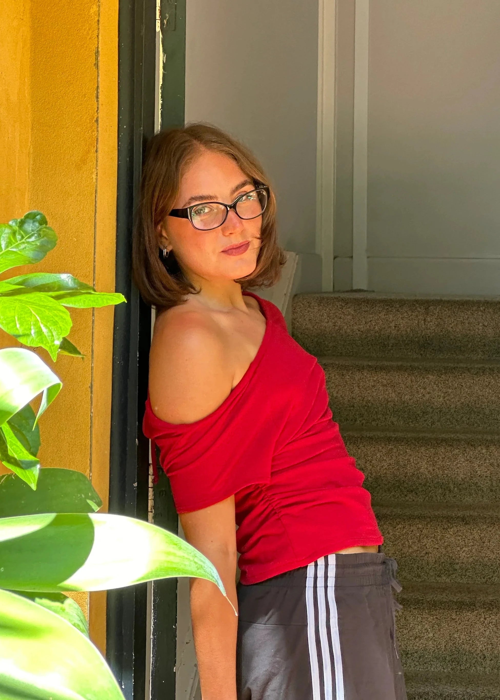 Woman in a red top and gray shorts leaning against a door with plants and stairs in the background