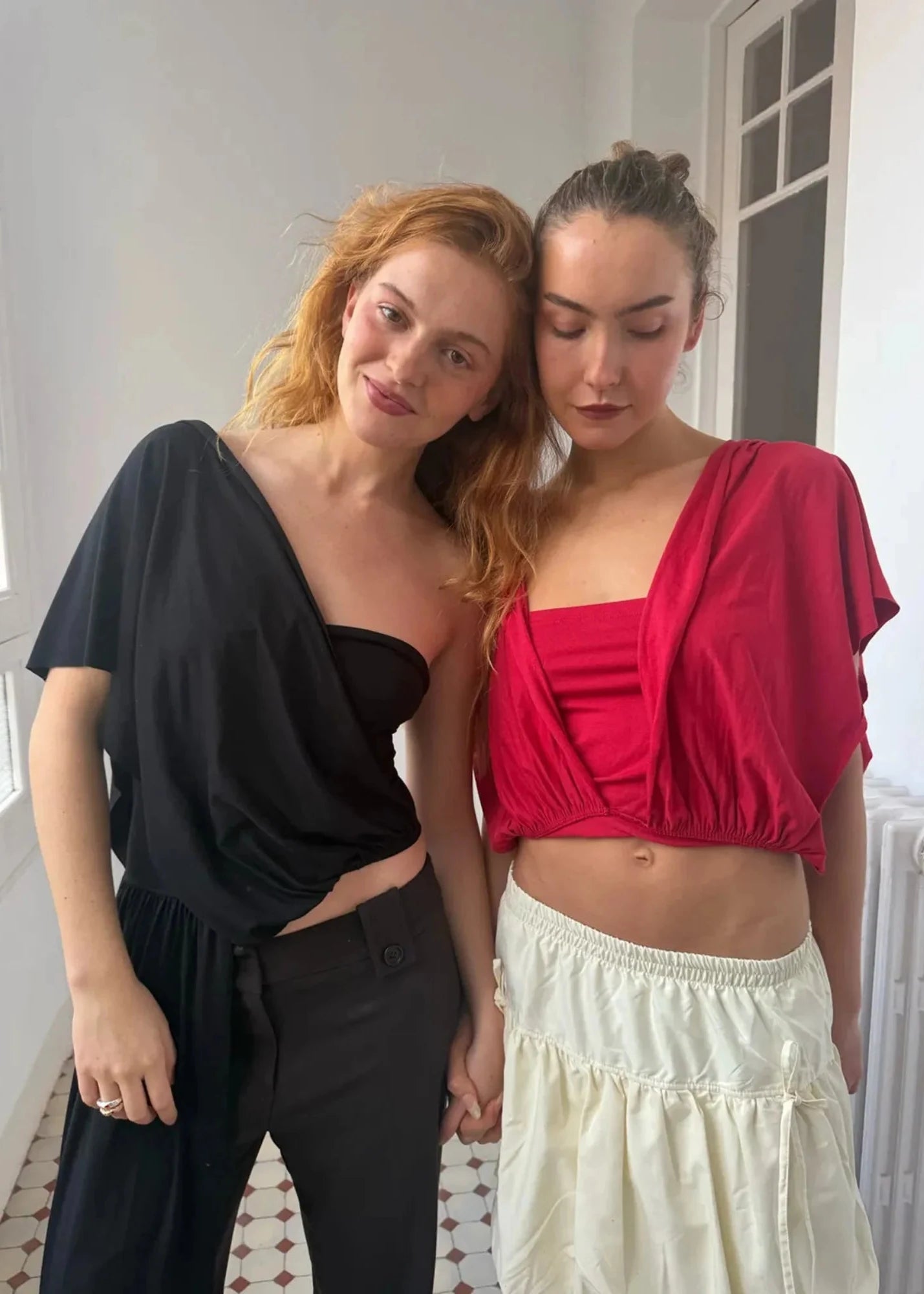 Two women standing close together indoors, one in a black outfit and the other in a red top and white skirt.