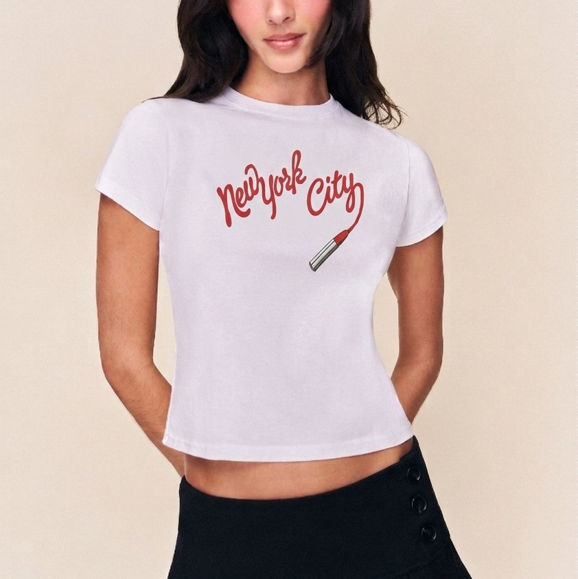 Woman wearing a white t-shirt with 'New York City' and a cigarette graphic, paired with a black pleated skirt on a beige background.