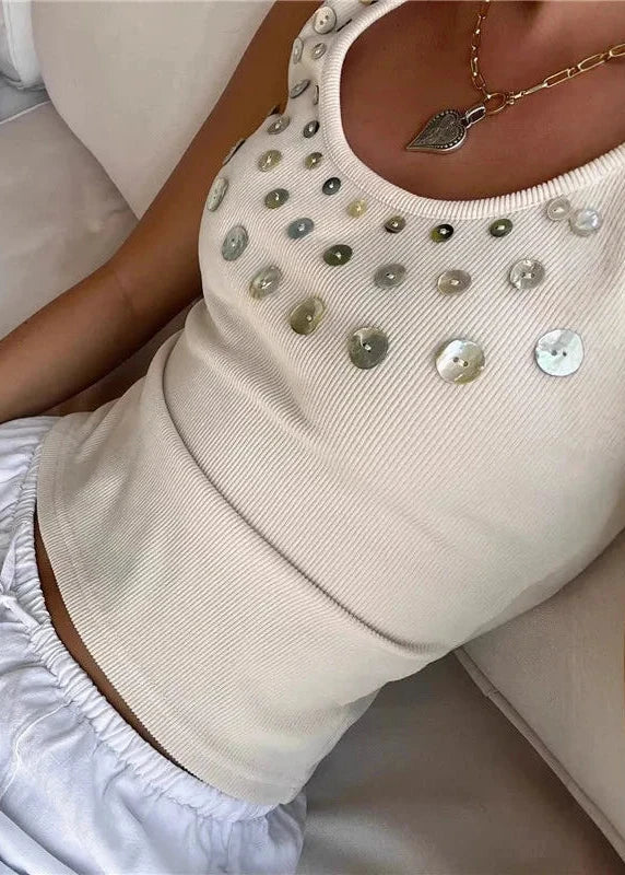 Person wearing a beige sleeveless top with sequins and white pants, sitting on a couch.