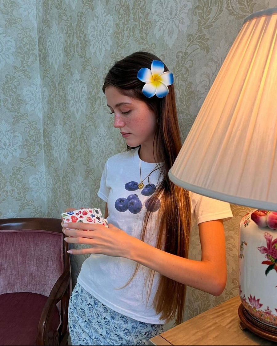 Young girl with a flower in her hair, holding a small object, sitting at a table with a lamp and chair in the background.