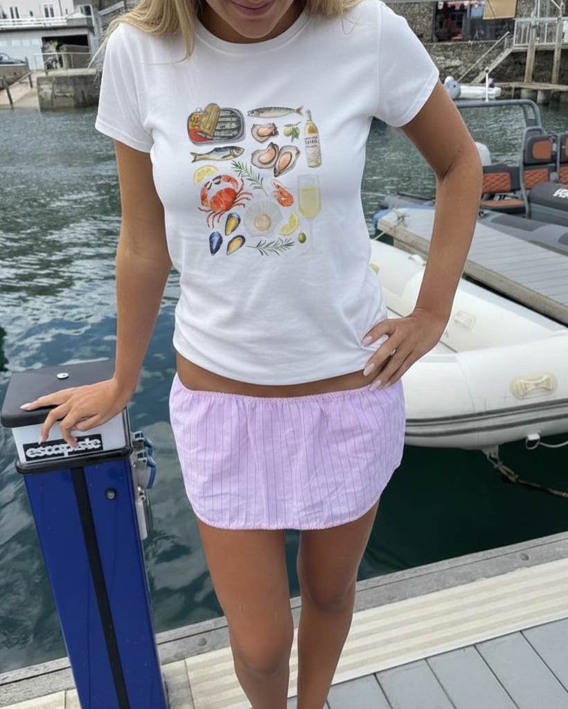 Person wearing a white t-shirt with a colorful design and a pink skirt standing on a dock with boats in the background.