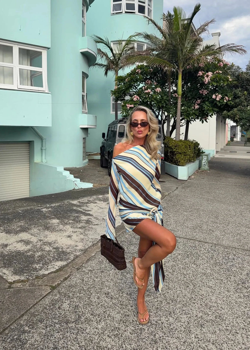 Woman in a striped dress posing on a street with a teal building and palm trees in the background