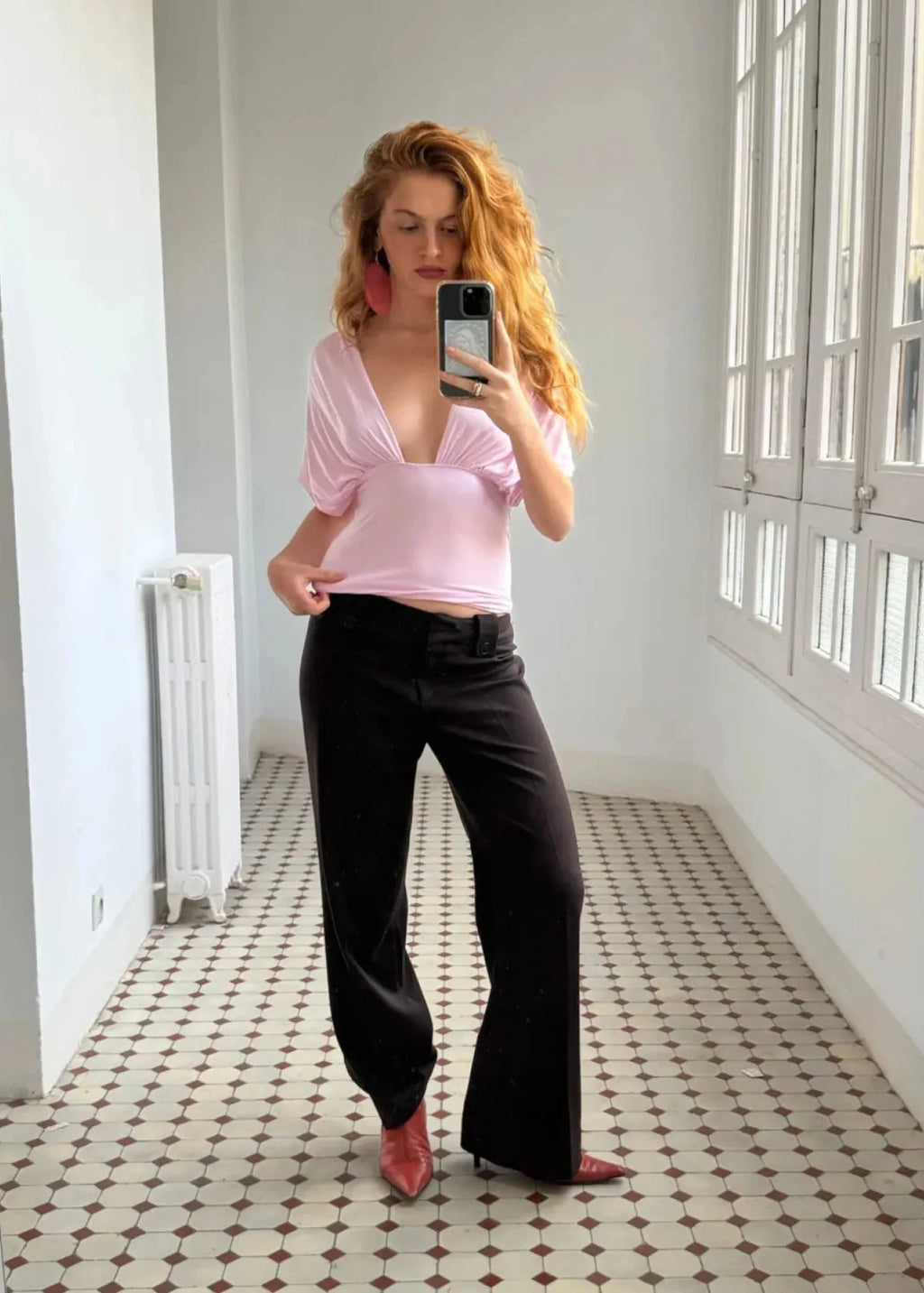 Woman taking a mirror selfie wearing a pink top and black pants in a room with white walls and patterned floor.