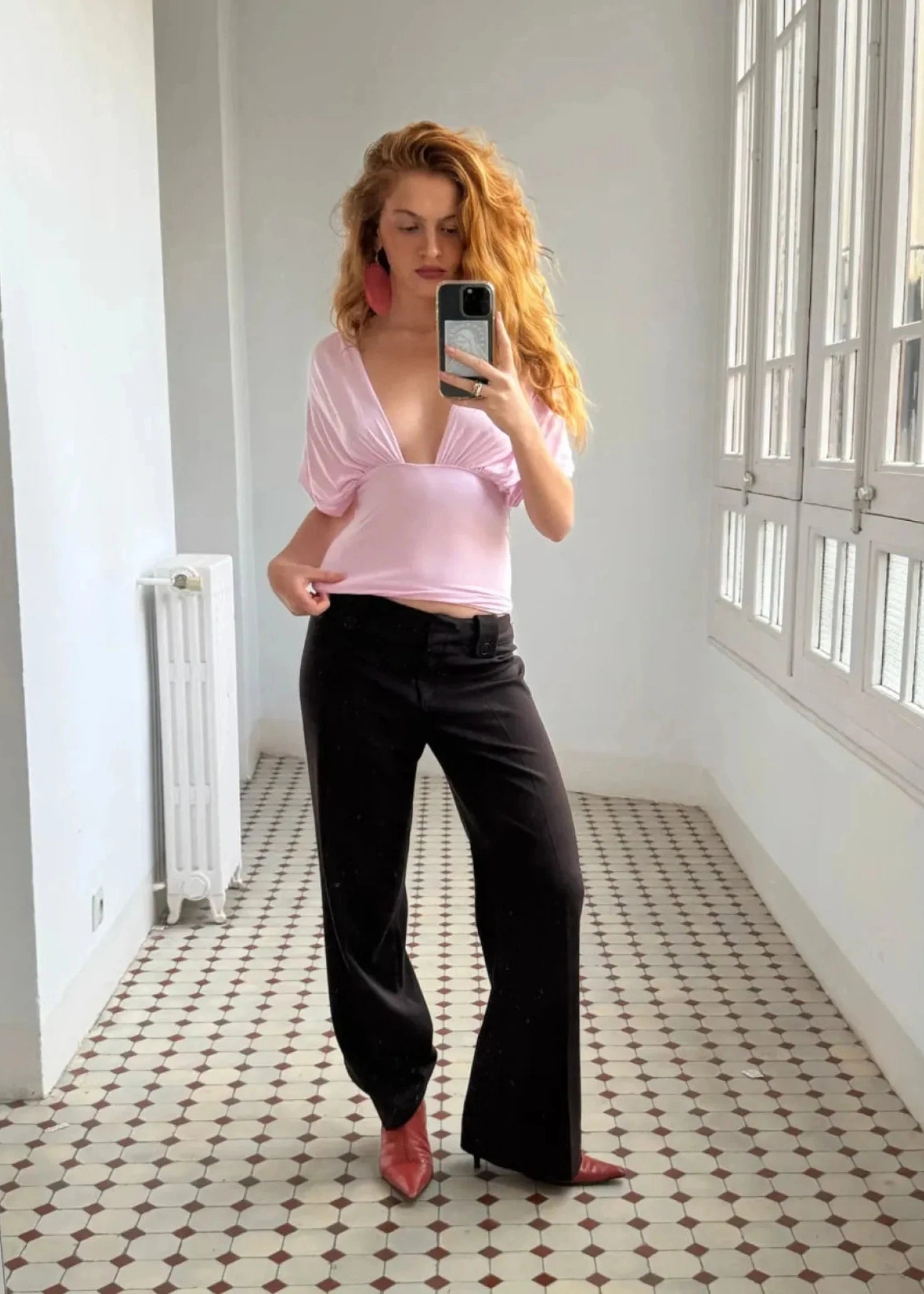 Woman taking a mirror selfie wearing a pink top and black pants in a room with white walls and patterned floor.