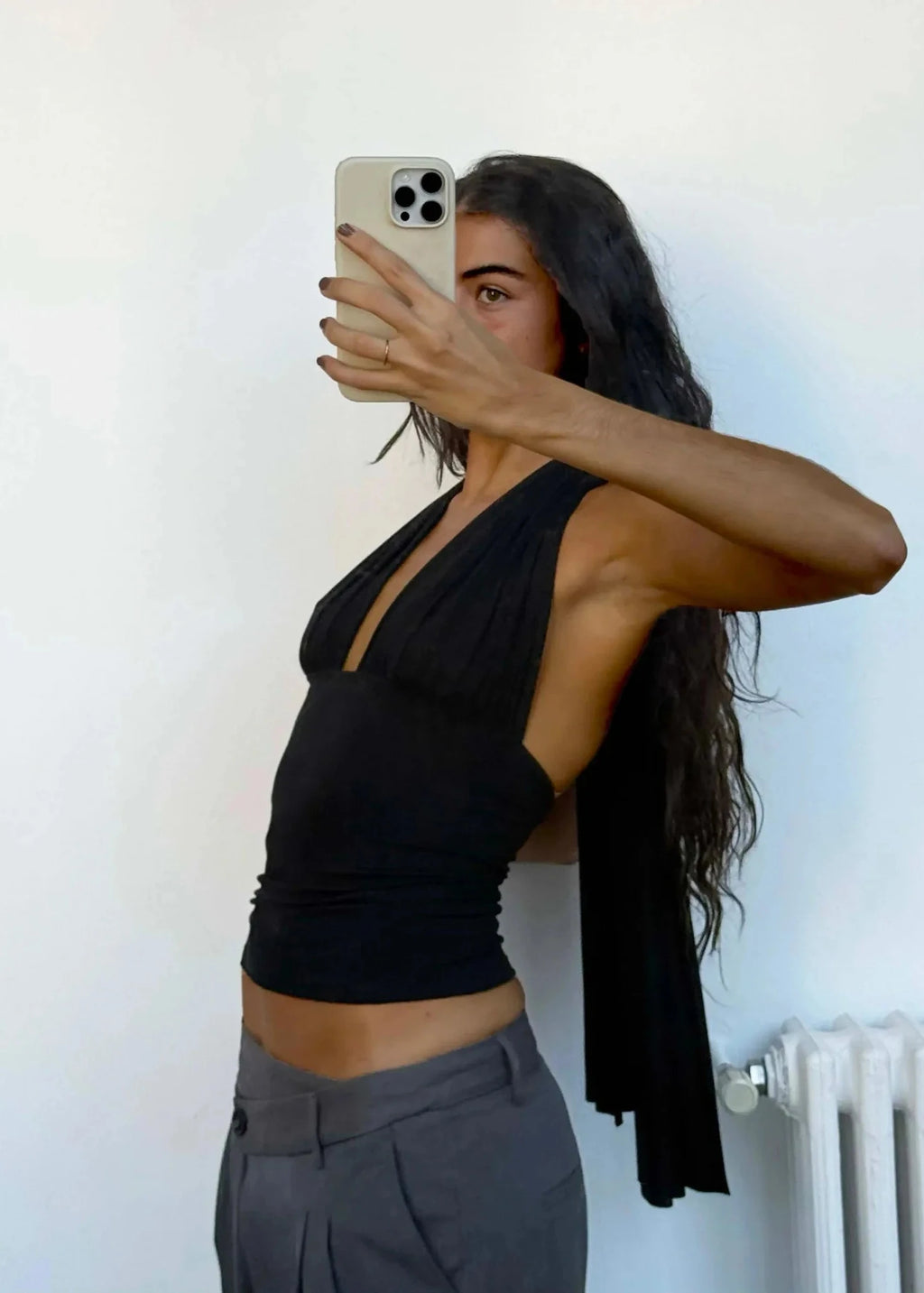 Woman taking a mirror selfie wearing a black top and gray pants against a white wall.