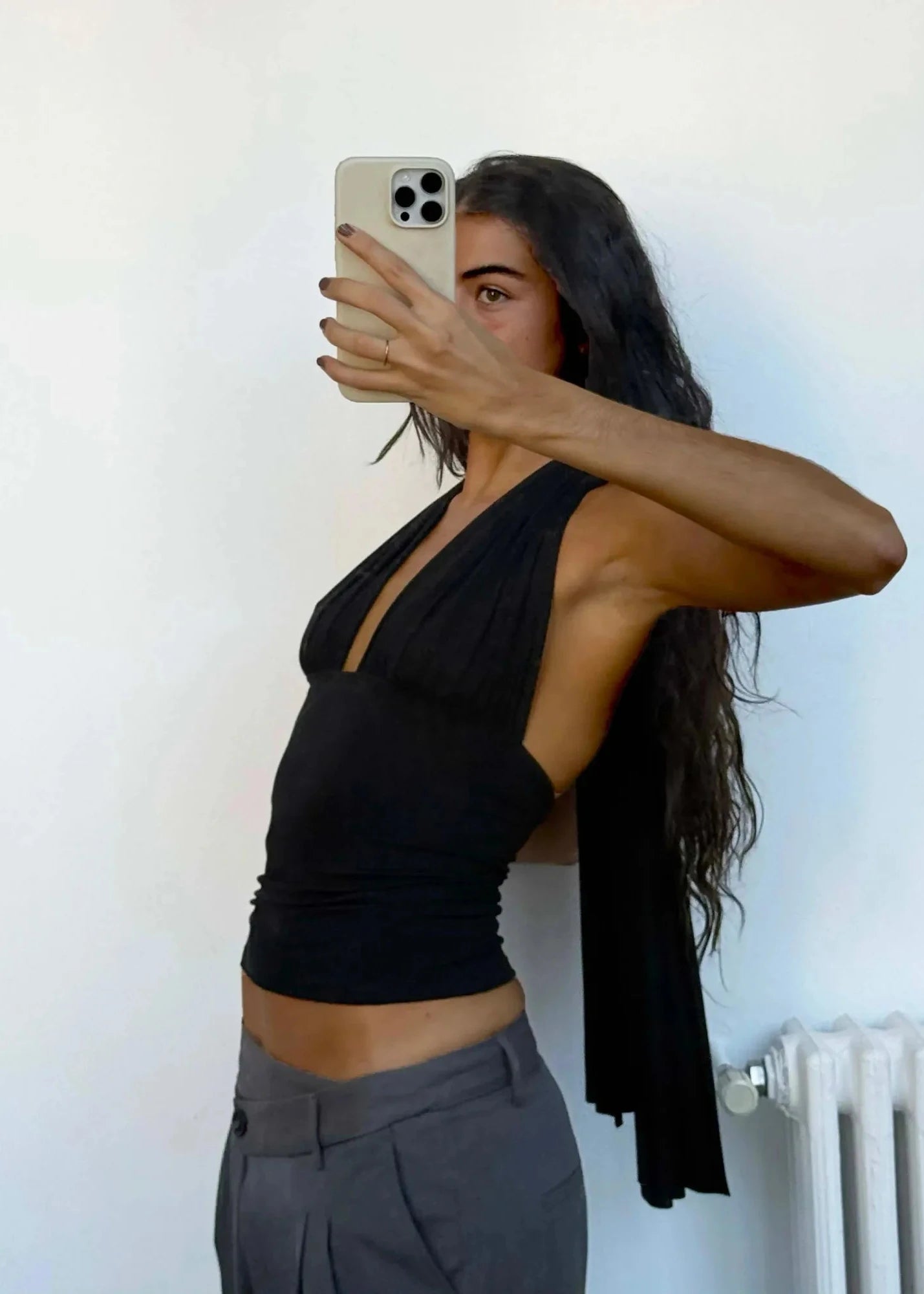 Woman taking a mirror selfie wearing a black top and gray pants against a white wall.