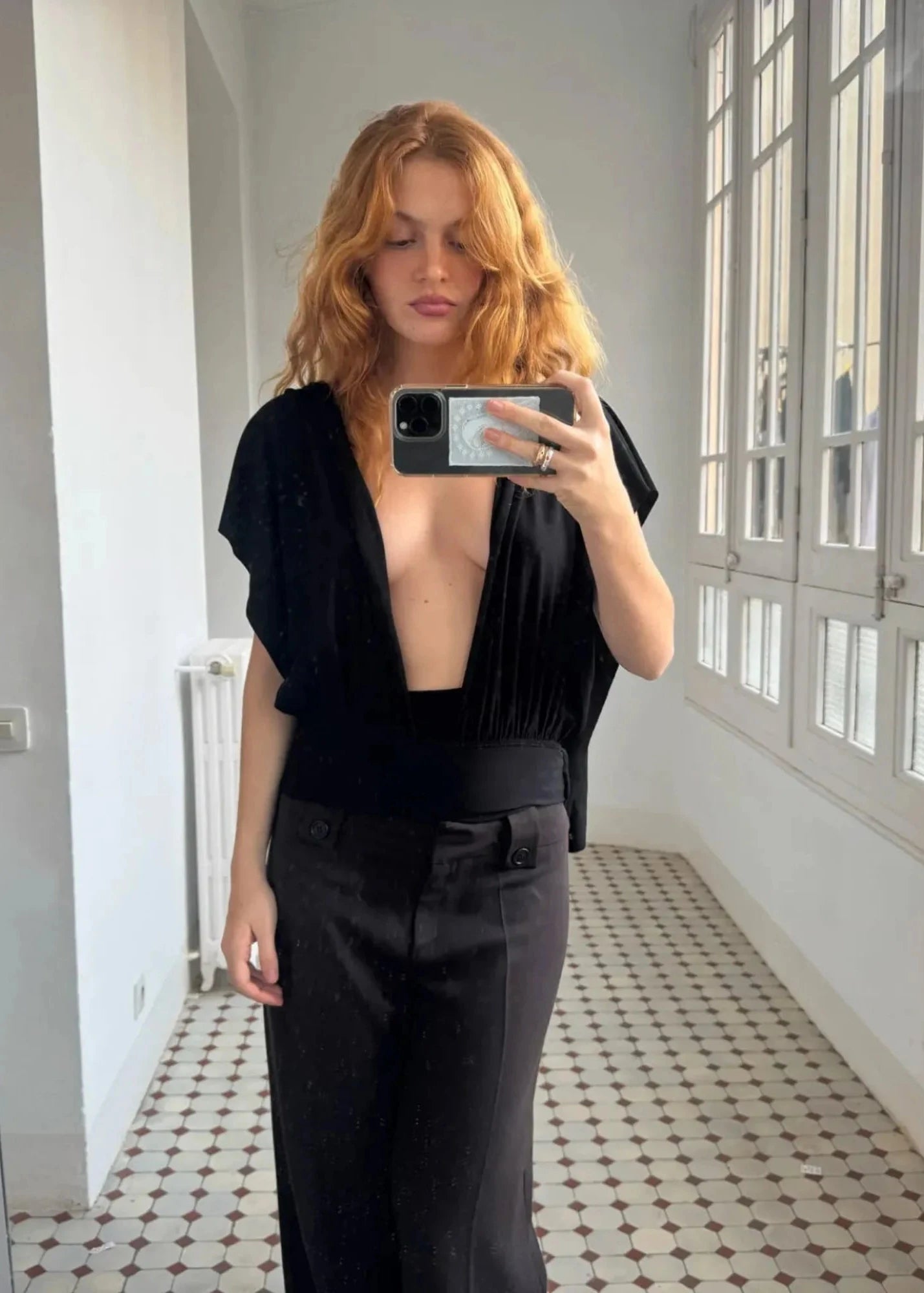 Person taking a mirror selfie wearing a black outfit in a room with white walls and tiled floor.