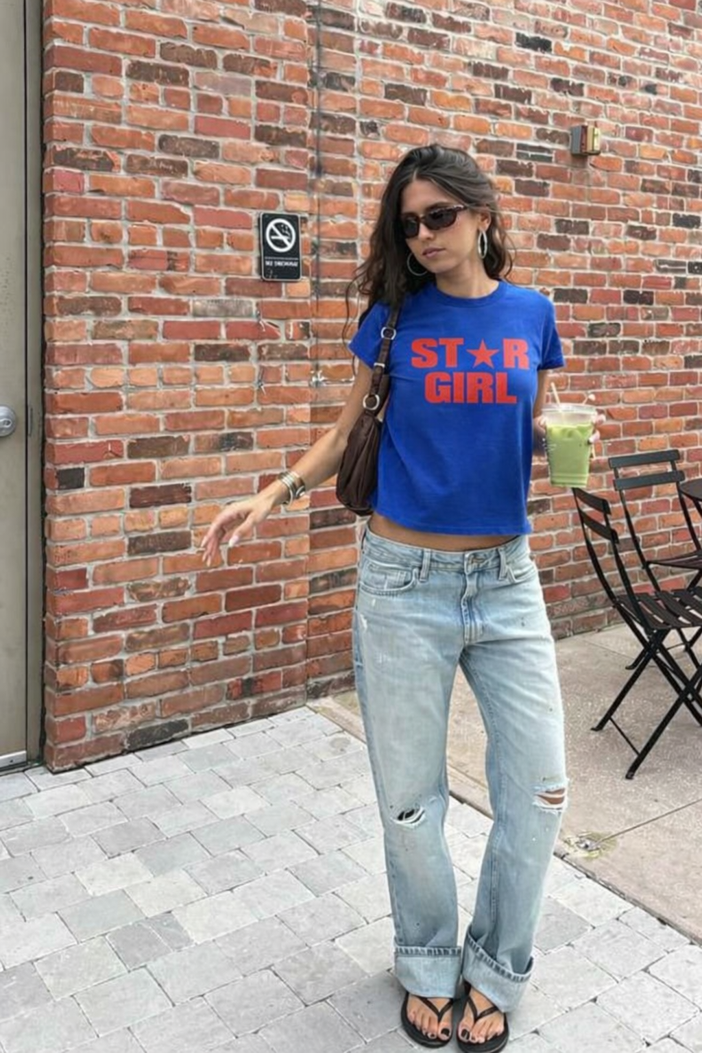 Woman wearing a blue 'STAR GIRL' t-shirt and light jeans standing against a brick wall.